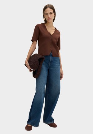 Kvinde iført brun, knappet top med korte ærmer, vide blå jeans, brune sko, holder en struktureret brun jakke, med langt, glat hår.