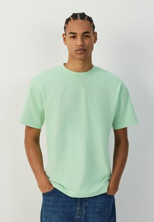 Jonge man met gevlochten haar, die een lichtgroen t-shirt en blauwe spijkerbroek draagt, staat met de handen in de zakken tegen een effen achtergrond.