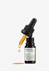 Bottiglia contagocce in vetro nero con etichetta verde, contenente siero arancione. Il contagocce è inclinato, con una goccia di siero che cade, su uno sfondo bianco.