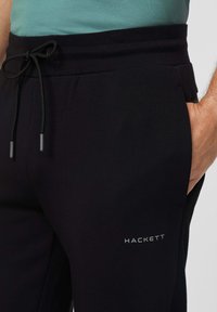 Schwarze Jogginghose mit einem Kordelzugbund, seitlichen Taschen und einem Logo mit silberner Druckfarbe auf dem linken Oberschenkel. Glatte Stofftextur.