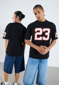 Svart t-shirt med kort ärm med ett vitt och rött nummer "23" tryckt på framsidan. Bärs med blå denimshorts, står rygg mot rygg.