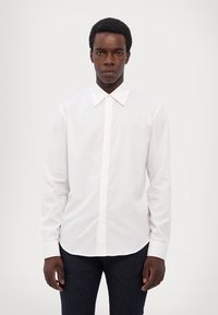 Λευκό πουκάμισο με μακριά μανίκια και κουμπιά, κλασικό γιακά, λείο ύφασμα και ίσια γραμμή; φοριέται με σκούρο τζιν.