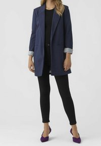 Granatowy długi blezer z podwiniętymi mankietami w paski, noszony na czarnym topie i czarnych spodniach. Stopy w fioletowych, teksturowanych szpilkach.