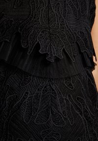 Tissu en dentelle noire avec une broderie florale complexe et un détail de bordure plissée au niveau du milieu du vêtement.