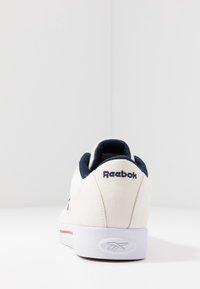 Zapatilla de lona blanca con un acento azul marino en el cuello del tobillo y una franja roja en la suela, que presenta un logo de Reebok en el talón.