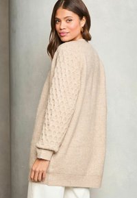 Beige svetr s texturovanými rukávy, vyrobený z měkkého pleteného materiálu, s uvolněným střihem a žebrovanými manžetami. Pozadí je v neutrální šedé barvě.