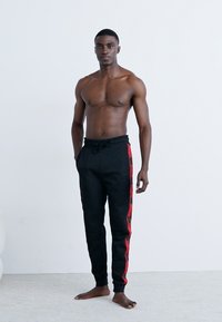 Zwarte sweatpants met rode zijstrepen, koordtailleband, taps toelopende pasvorm. Model staat topless tegen een neutrale achtergrond.