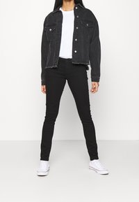 Chaqueta de mezclilla negra con dos bolsillos en el pecho y dobladillo deshilachado, combinada con jeans skinny negros y zapatillas de lona blancas. Camiseta blanca por debajo.