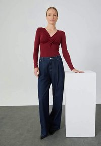 Burgundy long-sleeve top with a twisted front design, paired with high-waisted, wide-leg dark denim jeans. Smooth texture and tailored fit.