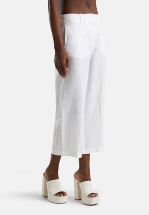 Modelo con pantalones cortos de lino blanco de pierna ancha y zapatos blancos de plataforma con tacón alto, puntera abierta y tipo slip-on sobre fondo liso.