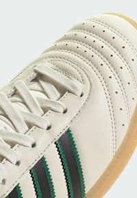 Biele tenisky s textúrovaným koženým zvrškom, s zelenými a čiernymi pruhmi, a gumovou podrážkou. Detaily zahŕňajú šité akcenty a perforácie.