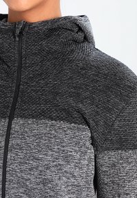 Sort og grå hættejakke med en tekstureret strikket mønster, med lynlås foran og en kontrastfarvet panel ved brystet.