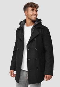 Svart ullblanding peacoat med dobbeltbrettet knappelukking, stor krage og kontrasterende grå hettefôr. Brukt over en hvit skjorte.
