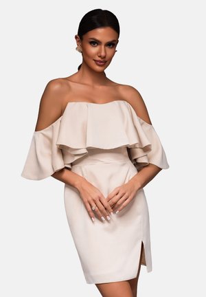 Beige mekko, jossa on olkaimia olkapäiltä, kerrokselliset liehukkeet, istuva vyötärö ja sivuaukko. Silkkinen kangas hienovaraisella kiillolla. Mallilla kädet ristissä.