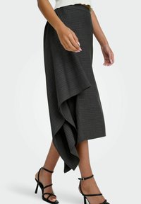 Vrouw in een donkergrijze asymmetrische midi-jurk met dunne verticale strepen en zwarte sandaaltjes met hoge hakken, aan het lopen.