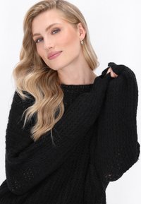 Pulover negru tricotat, cu o țesătură relaxată, mâneci largi și un decolteu rotund. Textura pare moale, cu o cădere casuală. Detalii subtile la manșete.