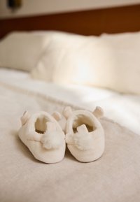 Crèmekleurige zachte babyslofjes met zachte pomponaccenten en oorachtige details; gladde textuur, afgeronde vorm, geplaatst op een licht stoffen oppervlak.