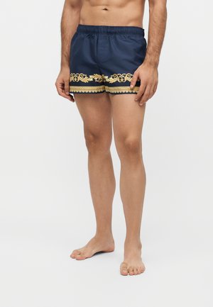 Torse et jambes d'un homme portant un short de bain bleu marine avec un imprimé décoratif doré le long de l'ourlet, debout pieds nus sur un fond clair.