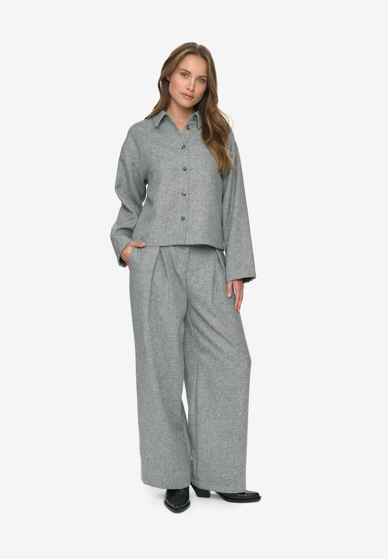 Femme debout avec une main dans la poche, portant une chemise grise à boutons et un pantalon large assorti, avec des bottines noires.