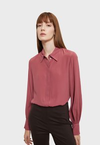 Ružová blúzka s gombíkovým golierom, dlhými rukávmi a detailmi na manžetách. Tkanina má hladkú textúru a jemný lesk. Skombinovaná s čiernymi nohavicami.