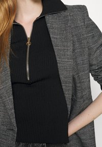 Blazer grigio a quadri con colletto a punta e dettagli sui bottoni, sovrapposto a un top nero a coste con chiusura a zip. Gli accessori includono hardware dorato.