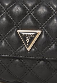 Borsa a tracolla in ecopelle nera trapuntata con una placca logata triangolare dorata con un punto interrogativo, texture in rilievo e dettagli cuciti.