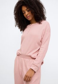 Ružový vrch s dlhými rukávmi z wafľovej pleteniny s tromi gombíkmi a rebrovanými manžetami, v kombinácii s voľnými nohavicami v rovnakej farbe, na neutrálnom pozadí.