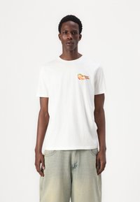 Λευκό βαμβακερό μπλουζάκι με έναν μικρό γραφικό πορτοκαλιού στην αριστερή πλευρά του στήθους. Η μπλούζα έχει χαλαρή εφαρμογή και στρογγυλή λαιμόκοψη.