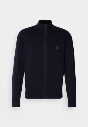 Marineblaue Zip-Up Strickjacke aus einem weichen Strickmaterial, mit hohem Kragen, langen Ärmeln und einem kleinen quadratischen Logo-Patch auf der Brust.