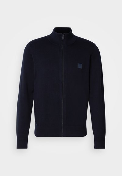 Cárdigan azul marino de cremallera fabricado en un material de punto suave, con un cuello alto, mangas largas y un pequeño parche de logo cuadrado en el pecho.