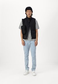 Zwarte puffer gilet over een grijze T-shirt, gecombineerd met lichtblauwe jeans. Het gilet heeft horizontale naden en een zijtas. Witte sneakers.
