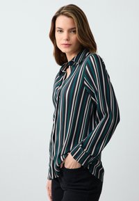 Koszula w paski w kolorach czarnym, zielonym i białym; długie rękawy; luźny krój; subtelny połysk; posiada kołnierzyk i mankiety.