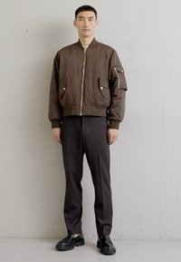Geacă bomber maro cu fermoar frontal și două buzunare laterale; manșete și tiv ribbate; purtată peste pantaloni gri închis și pantofi negri.