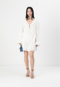 Λευκό φόρεμα με floral σχέδιο, με φουσκωτά μανίκια, λαιμόκοψη με κορδόνι και πολλαπλά στρώματα. Συνδυασμένο με σανδάλια με λουριά και μπλε τσάντα.