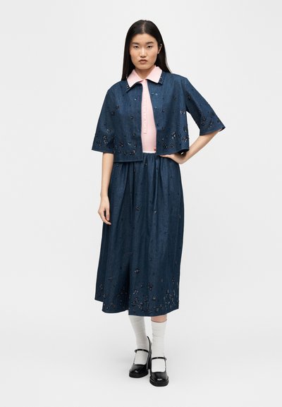 Femme aux longs cheveux bruns portant une jupe bleue brodée de fleurs et une veste courte assortie sur une chemise rose à col, des chaussures noires et des chaussettes blanches.