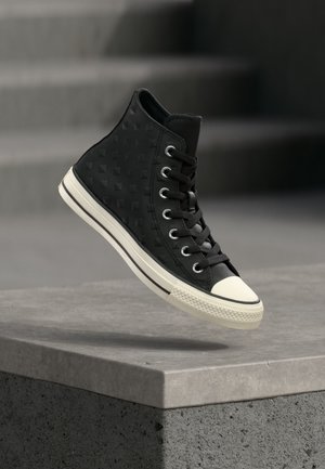 Zapatilla negra de caña alta con lateral texturizado, puntera de goma blanca y suela blanca, capturada en el aire sobre escalones de hormigón.