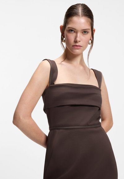 Jeune femme aux cheveux bruns portant une robe marron foncé sans manches, avec des boucles d'oreilles anneaux dorés, posant devant un fond blanc uni.