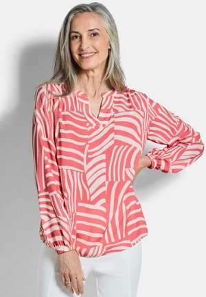 Frau mit grauen Haaren, die eine pink-weiß gestreifte Bluse mit abstraktem Muster und weiße Hosen trägt, lächelnd mit der Hand in der Hüfte.