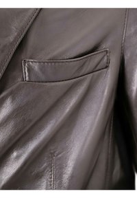 Nahaufnahme einer braunen Lederjacke mit einer glatten Textur, einer kleinen Brusttasche und sichtbaren Nähten entlang der Kanten.