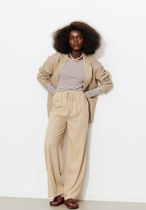 Femme aux cheveux bouclés et volumineux, portant un blazer beige à fines rayures, une chemise rayée, un pantalon large beige, des sandales et un collier de perles, debout les mains sur les hanches.