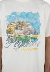 T-shirt en coton blanc présentant une impression aquarelle colorée d'une ville côtière et le texte "Pegador Scent of Summer" en argent et jaune.