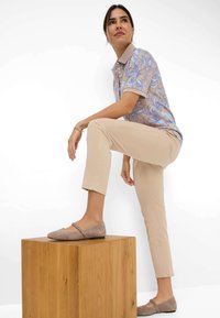 Hellbeige Hose und ein blaues Polohemd mit floralem Muster und beigem Kragen. Graue flache Schuhe mit Rüschenakzenten. Steht auf einem Holzblock.
