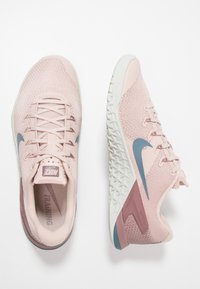 Zapatillas de entrenamiento Nike de color rosa claro con parte superior de malla transpirable, acentos azules, suela texturizada y detalle en el talón en un acabado gris claro.