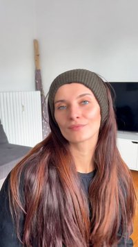 Păr lung, ondulat, de culoare brunetă cu reflexe roșcate; poartă o bandă de cap din tricot de culoare verde închis și o bluză neagră cu mânecă lungă. Iluminare moale, fundal neutru.