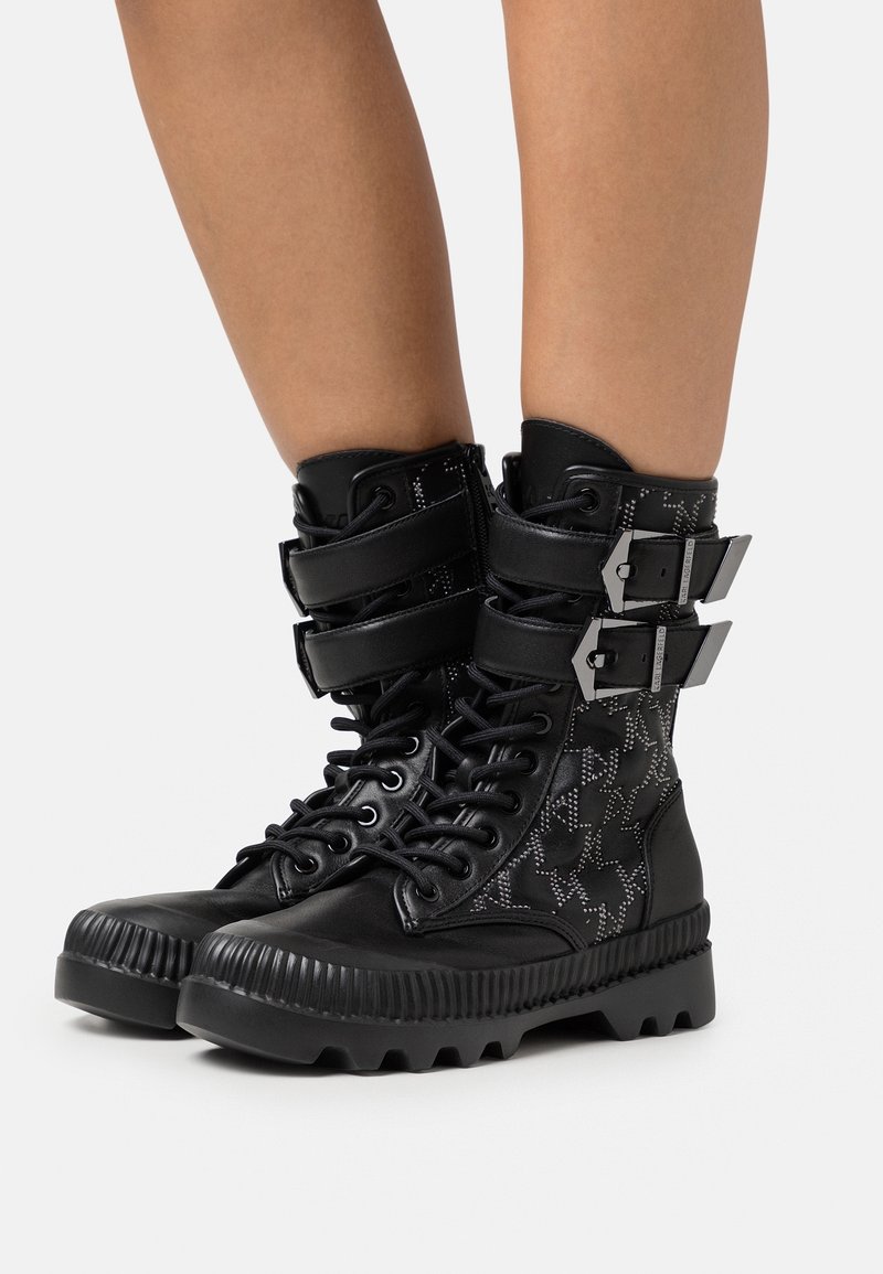 Bottes montantes en cuir noir avec lacets, doubles bandes velcro, détails avec motif d'étoiles, et semelles en caoutchouc épaisses sur jambes nues.