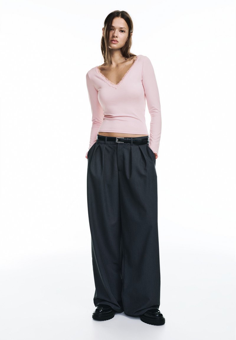 Bluză roz cu mâneci lungi și decolteu din dantelă, asortată cu pantaloni evazați gri închis și pantofi negri lucioși, prevăzuți cu o curea.