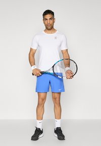Tennis player wearing a white, short-sleeve shirt and blue shorts. He holds a black tennis racket and wears black athletic shoes with white socks.