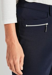 Marineblaues Paar Hosen mit glatter Textur, ausgestattet mit einer seitlichen Reißverschlusstasche. Nahaufnahme einer Hand, die auf dem Stoff ruht, und die Details und Nähte zeigt.