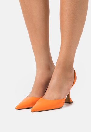 Pieds portant des escarpins à bride arrière orange vif à bouts pointus avec des talons blocs bas et inclinés sur un fond blanc.