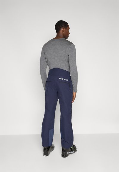 Σκούρο μπλε παντελόνι για σκι με εφαρμοστή μέση, υφασμάτινη υφή, τσέπες με φερμουάρ και λογότυπο στο πίσω μέρος. Φοριούνται με μαύρα παπούτσια σκι.